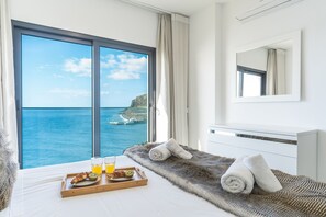 Une chambre d’hôtel dotée d’une grande fenêtre donnant sur la mer, d’un lit recouvert d’une couverture moelleuse et agrémenté de serviettes, ainsi que d’un plateau de petit-déjeuner comprenant des croissants, des kiwis et du jus d’orange.