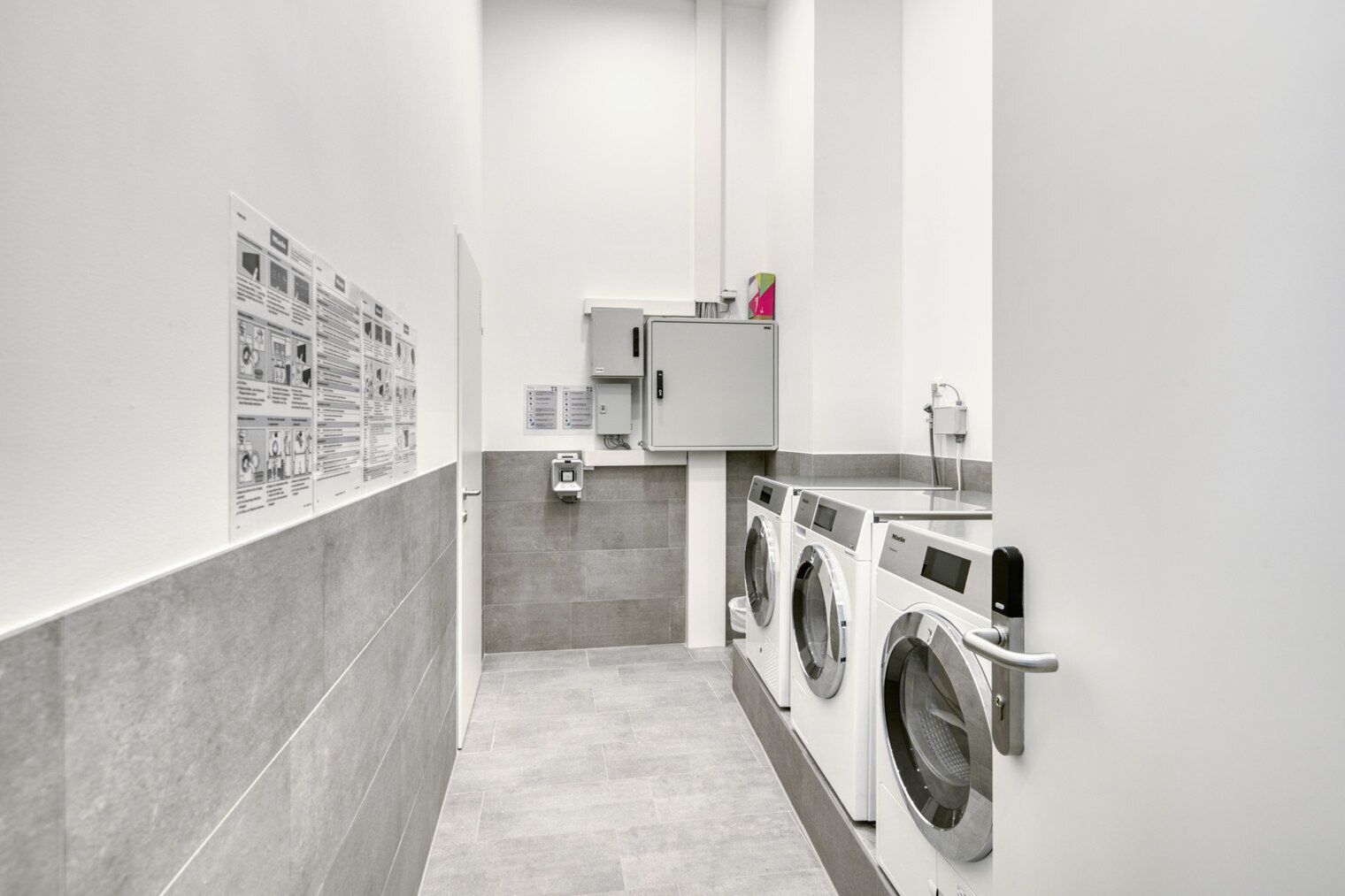 A modern laundry facility with two front-loading washing machines, a utility cabinet, and informational posters on the wall.