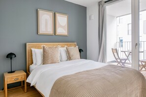 Quarto com cabeceira de madeira, cama com roupa de cama branca, duas obras de arte abstrata emolduradas, mesa de cabeceira com luminária e uma varanda com cadeiras de madeira.