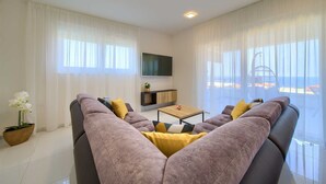Un salon moderne avec un grand canapé, une table basse et un téléviseur. On y trouve une plante en pot et, grâce à de grandes fenêtres, une vue sur la mer.