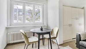 Een kamer met een witte radiator, een ronde tafel met twee stoelen, een raam met uitzicht op bomen, en een witte garderobe.