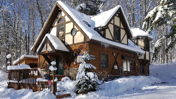 Una cabaña de madera con techo cubierto de nieve, rodeada por un paisaje nevado y árboles.