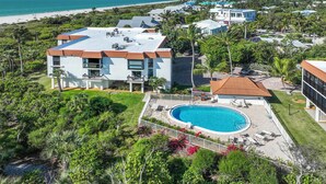 A modern two-story building with a rooftop pool, surrounded by lush greenery and a view of the beach.
