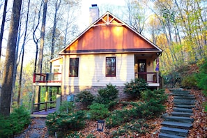 Une cabane en bois dotée d’un balcon, entourée d’arbres, et un escalier en pierre menant à l’entrée.