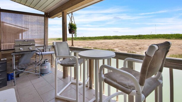 A covered outdoor area with a grill, chairs, and a table, overlooking a sandy landscape.