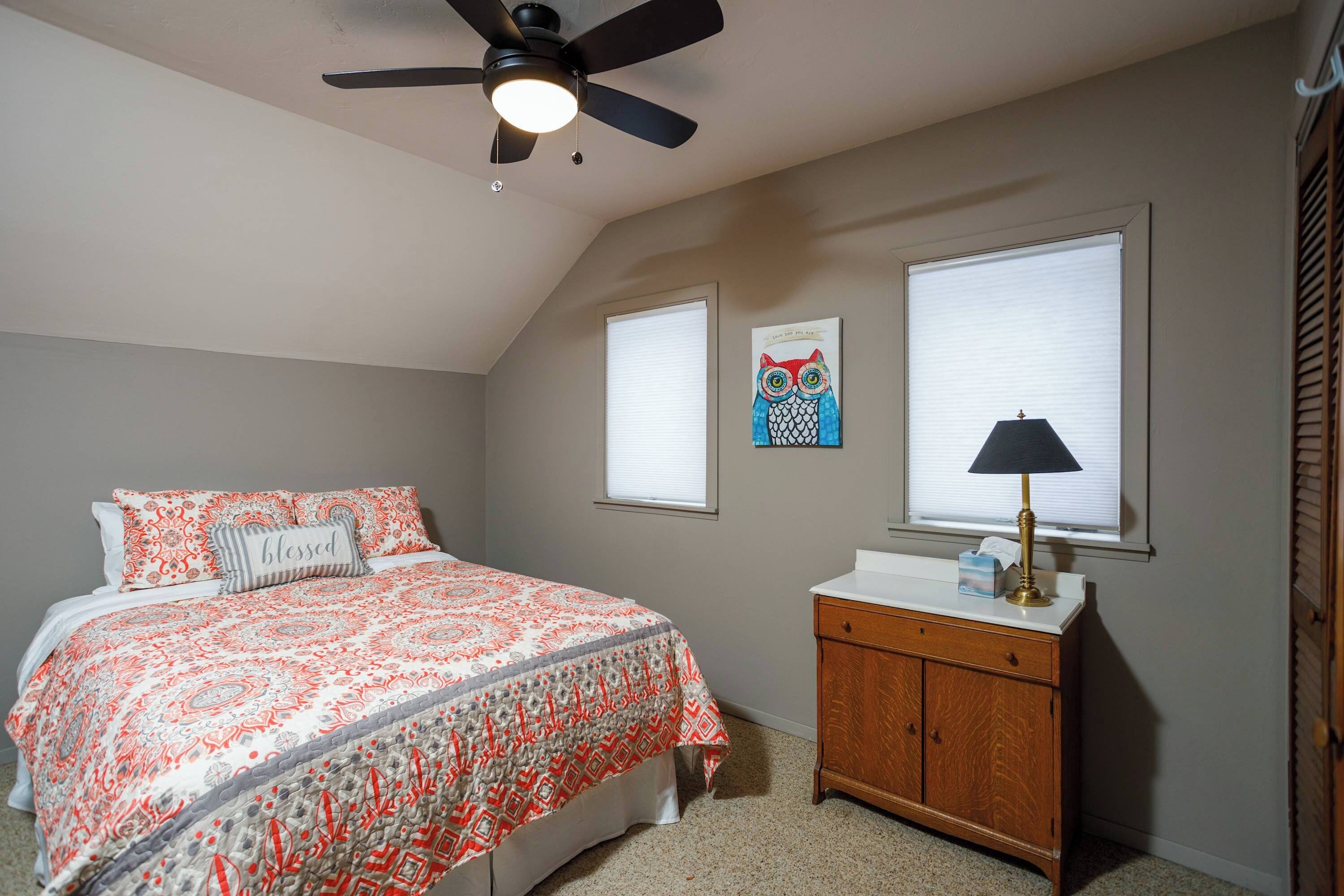 A bedroom with a bed, a ceiling fan, a nightstand, and two windows.
