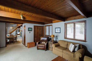 A spacious living room with a high wooden ceiling, a leather armchair, a beige sofa, and a dining area with a wooden table and chairs.