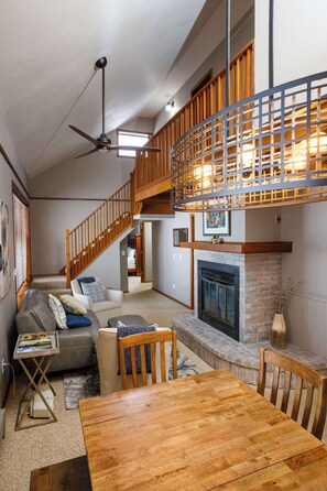 Un salón amplio con techo alto, chimenea, mesa de comedor y escalera que conduce a un nivel superior.