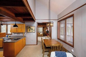 A modern kitchen with wooden cabinets, a dining area with a wooden table and chairs, and a large window with blinds. - 185' Waterfront Sand Bay Owl's Nest 4BR sleeps 9 (Sturgeon Bay)