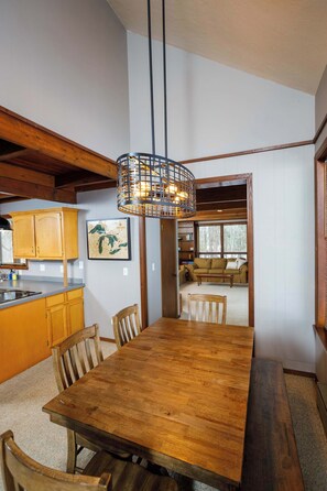 A wooden dining table with chairs, a hanging light fixture, wooden cabinets, and a view into a living area.