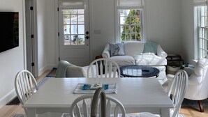 A dining area with a white table, chairs, and a chandelier. There is a sofa with a blue cushion and a window with a view of trees. - The Rose Cottage of Sandwich Village (Sandwich)