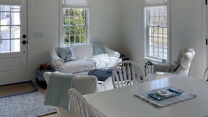 A bright, white room with a wooden ceiling, a white sofa with blue cushions, a dining table with chairs, and large windows with white blinds. - The Rose Cottage of Sandwich Village (Sandwich)