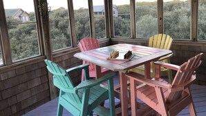 Um alpendre coberto com uma mesa de madeira e cadeiras coloridas, grandes janelas com vista para árvores e casas, e luzes de Natal.