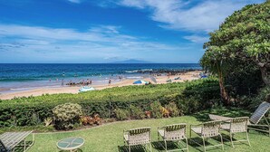 A beach view with lounge chairs and a table, a clear sky, and a distant mountain.
