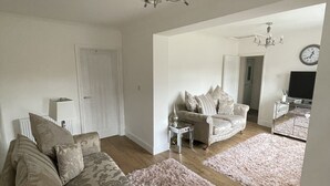 A spacious living room with a large sofa, a coffee table, and a chandelier. There is a door leading to another room and a clock on the wall.