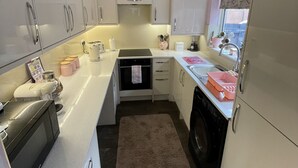A compact kitchen with white cabinets, a built-in oven, a washing machine, and a window with a heart-patterned blind.