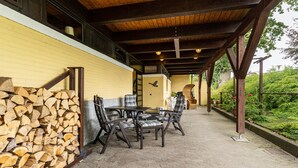 An outdoor covered patio with a wooden ceiling, a table with chairs, and a stack of firewood against a wall.