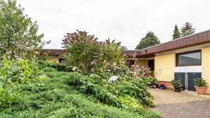 A garden with various plants and flowers, a paved walkway, and a building with a brown roof and yellow walls.
