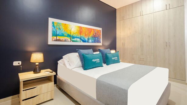 A hotel room with a bed, a nightstand, a lamp, a painting, and a wardrobe. - Montepinares Apartaestudios (Montería)