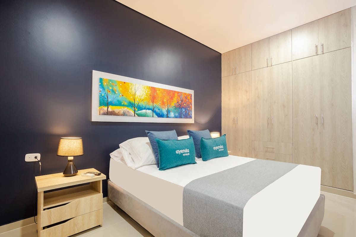 Hotellihuone, jossa on sänky, yöpöytä, lamppu, taulu ja vaatekaappi.