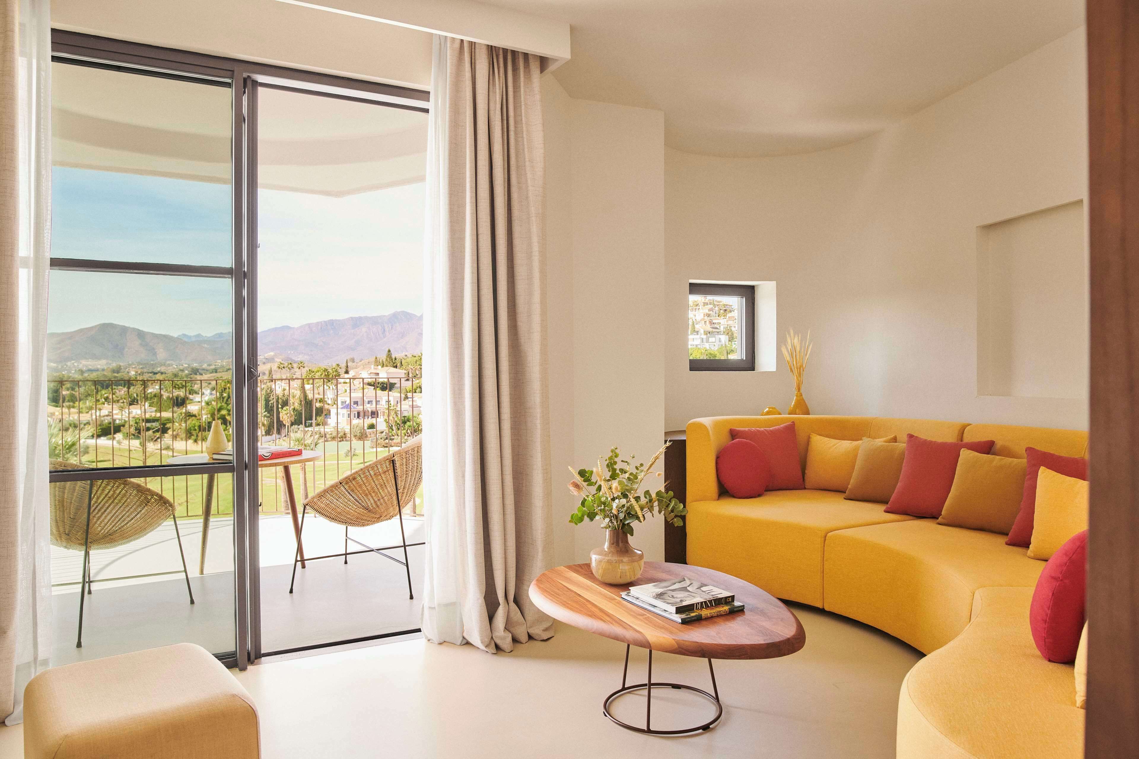 Una sala de estar moderna con un sofá amarillo, una mesa de centro de madera y vistas a un paisaje urbano a través de grandes puertas de cristal.