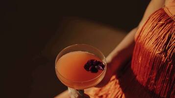 A cocktail in a martini glass with a garnish, placed on a surface next to a person wearing a red fringe garment.