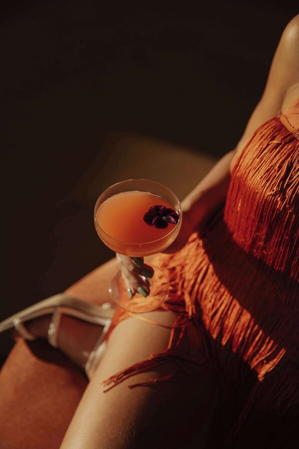 A cocktail in a martini glass with a garnish, placed on a surface next to a person wearing a red fringe garment. - La Zambra Resort Mijas - Marbella The Unbound Collection By Hyatt (Mijas)