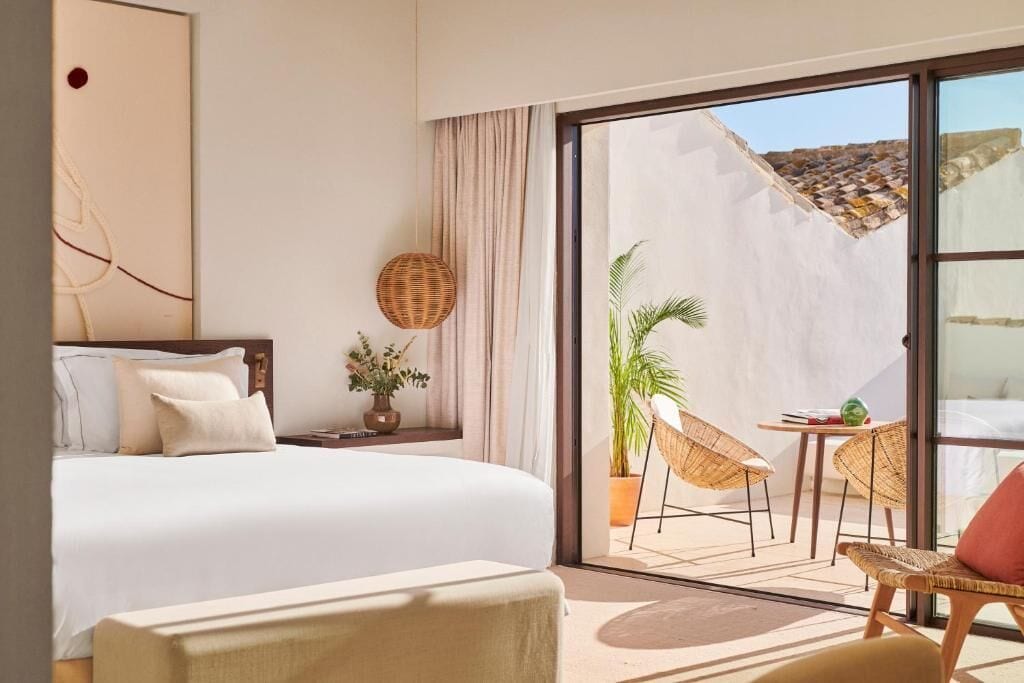 A hotel room with a bed, a small table with chairs, a potted plant, and a view of the outside.