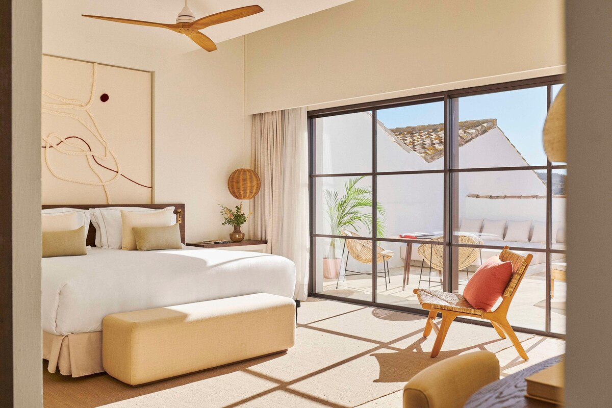 Un dormitorio espacioso con una cama grande, un ventilador de techo de madera y vistas al exterior a través de un amplio ventanal.