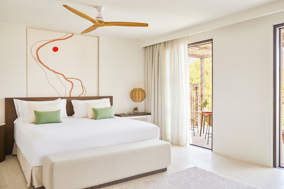 Habitación de hotel moderna con una cama grande, ventilador de techo, vista del patio exterior y un cuadro abstracto de gran tamaño en la pared.