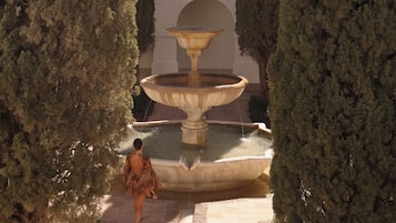 A woman walking towards a multi-tiered fountain in a courtyard surrounded by tall trees.
