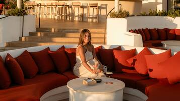 Un área de estar moderna al aire libre con un sofá blanco curvo y cojines rojos, una mesa de centro blanca redonda y una zona de bar al fondo.
