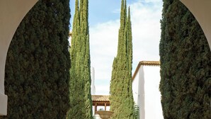Un patio con una fuente central, rodeado de cipreses podados y un edificio blanco de techo con tejas.
