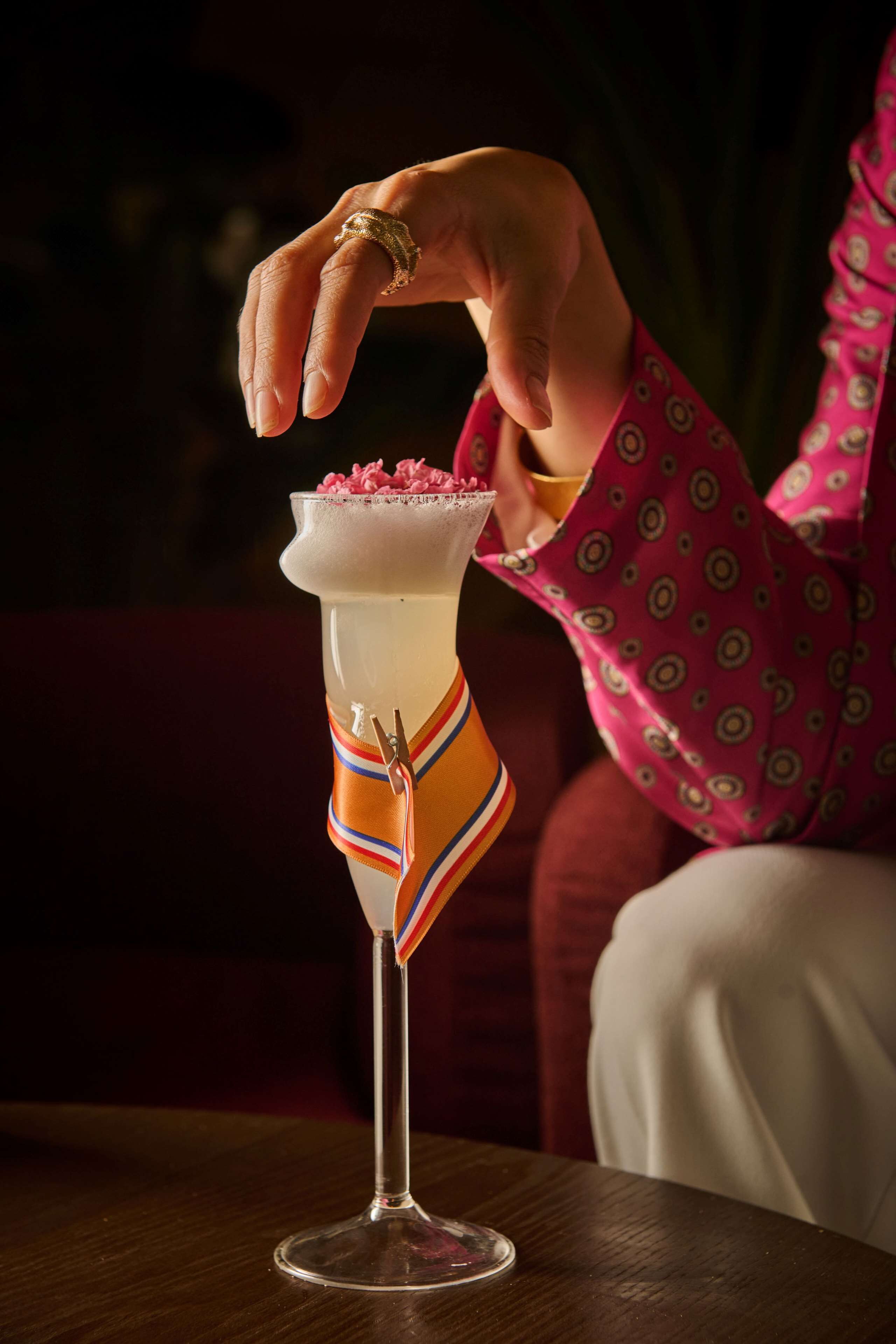 En cocktail i ett högt glas, dekorerad med ett färgglatt band, en hand med en ring och en person i en mönstrad, rosa skjorta.