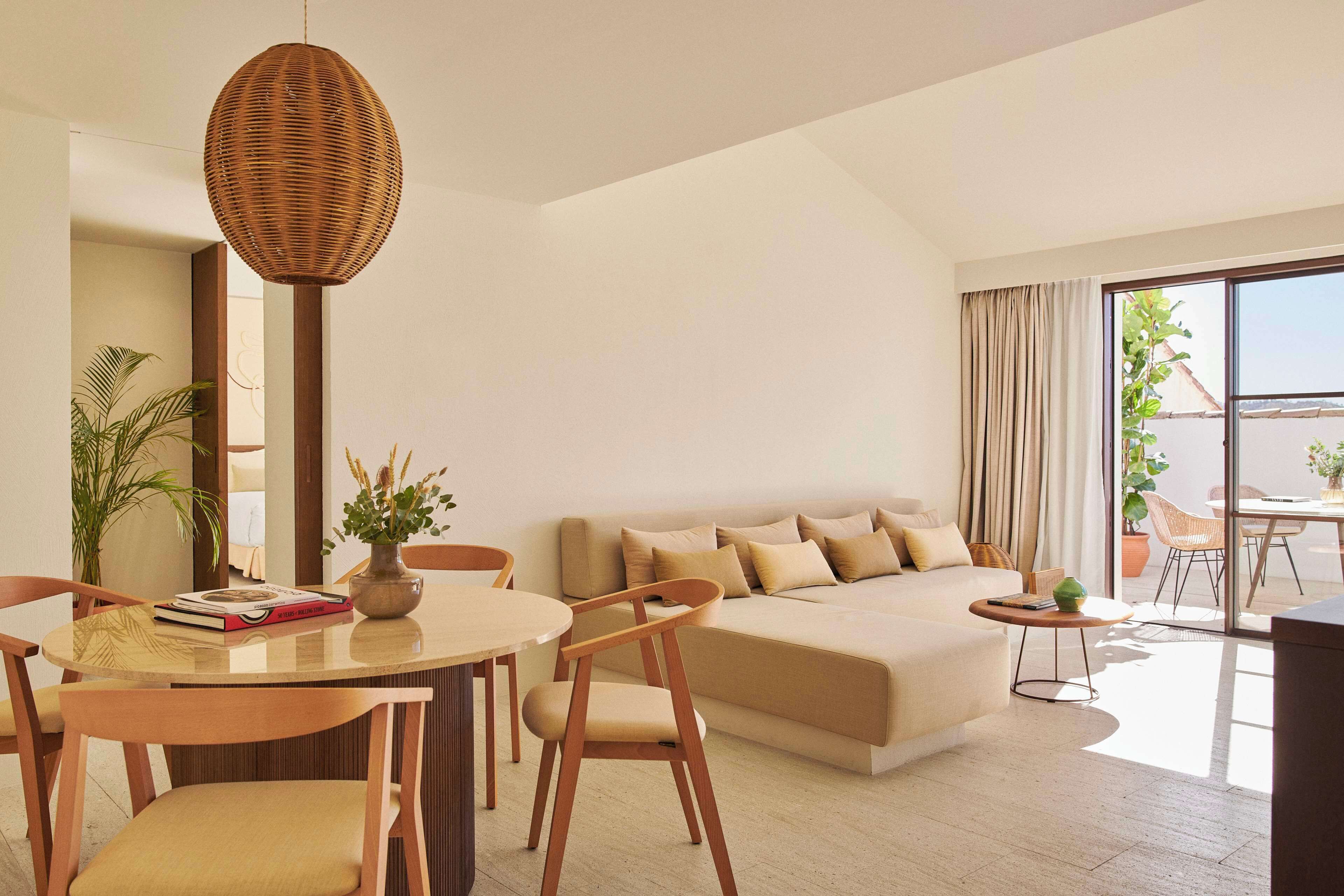A modern hotel room with a large bed, a dining area with a round table and chairs, a wicker pendant light, and a view of the outdoors through large windows.