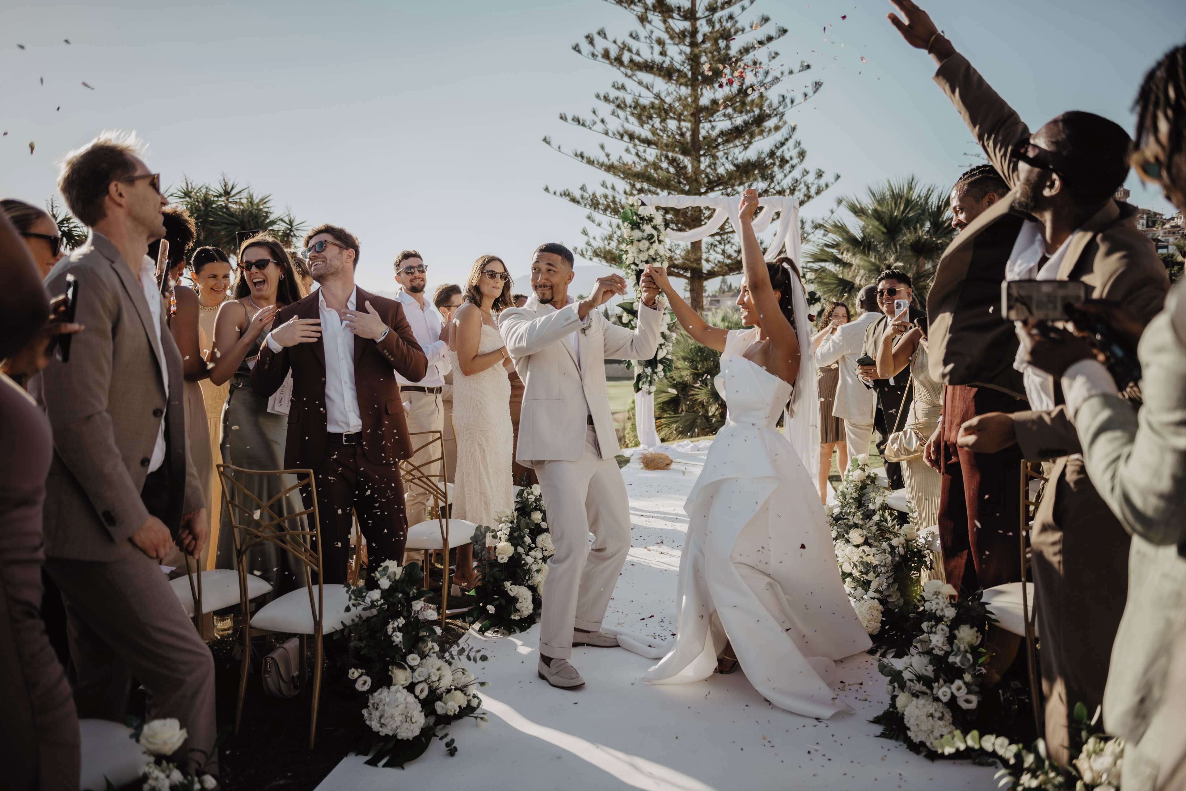 Una ceremonia de boda con invitados sentados en sillas, una novia con vestido blanco y un novio en traje.