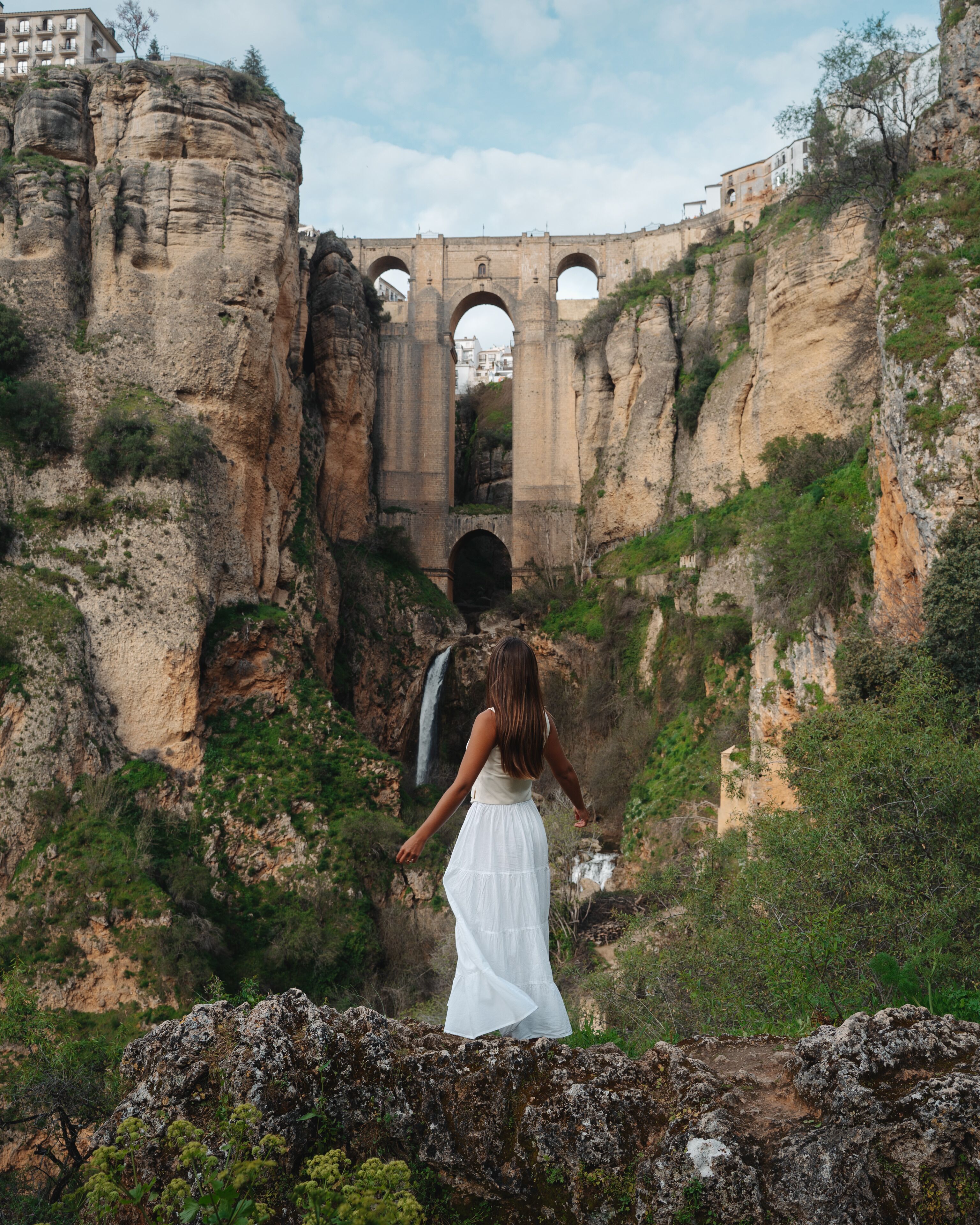 Une femme vêtue d’une robe blanche se tient devant un pont de pierre à arcades, surplombant un paysage rocheux parsemé de verdure.