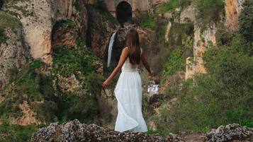 A woman in a white dress standing in front of a stone bridge with arches, overlooking a rocky landscape with greenery.