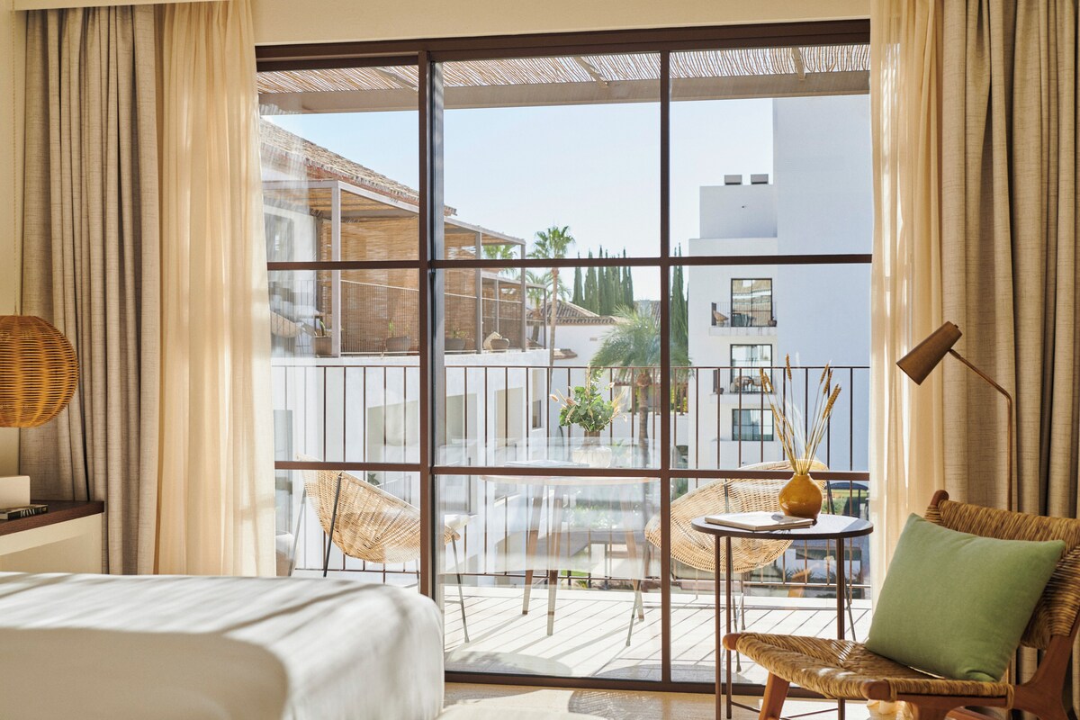 A hotel room with a large window, a bed, a wicker chair, a small table with a vase, and a balcony with a view of buildings and palm trees.
