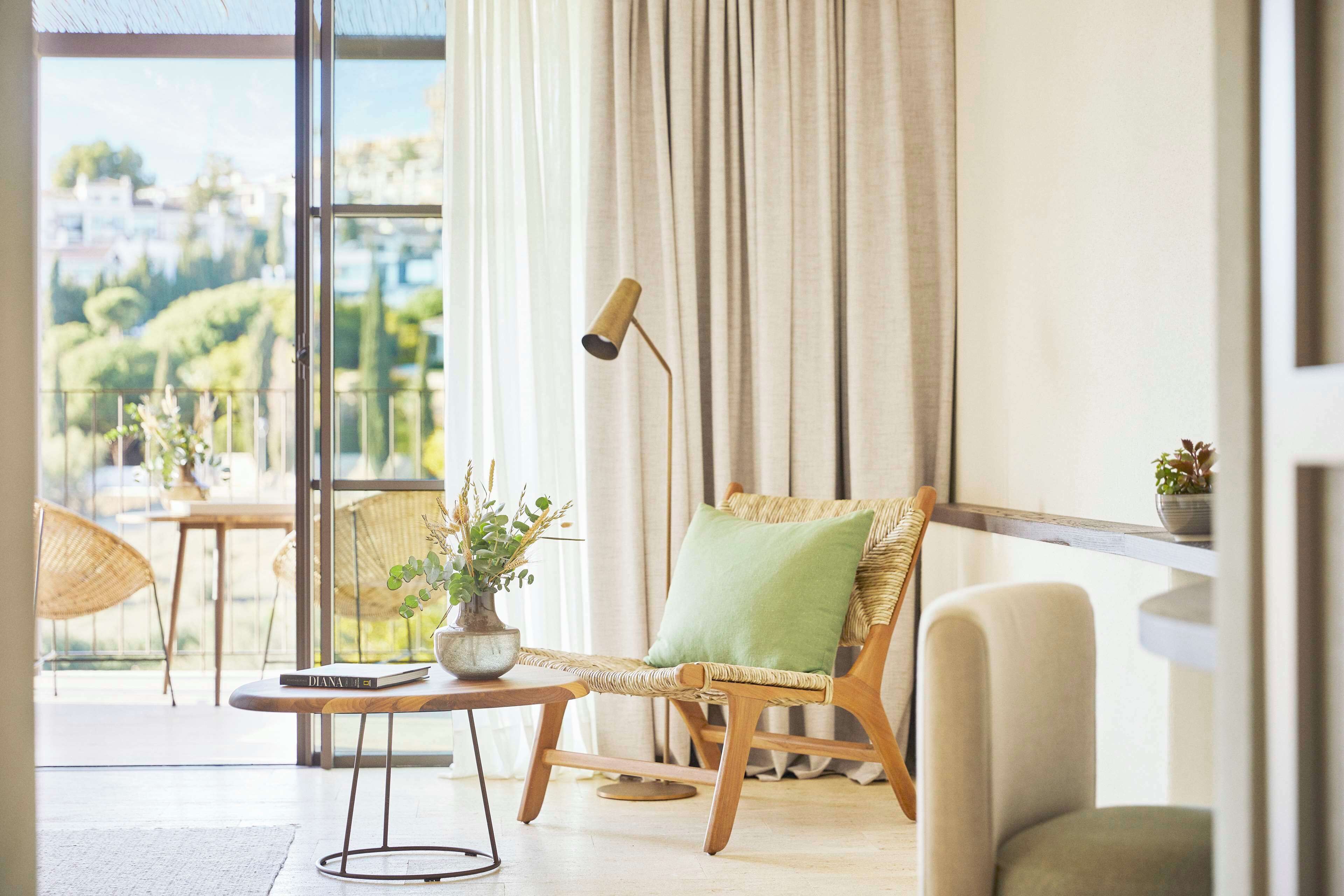 Una sala moderna con una silla de mimbre, un cojín verde, una mesita con un libro y una planta, y vistas a zonas verdes al exterior.