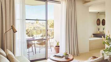 Una sala de estar moderna con sofá, una mesita redonda con un libro y una planta, un balcón con una silla y una mesa, y una zona de cocina con un fregadero y adornos.