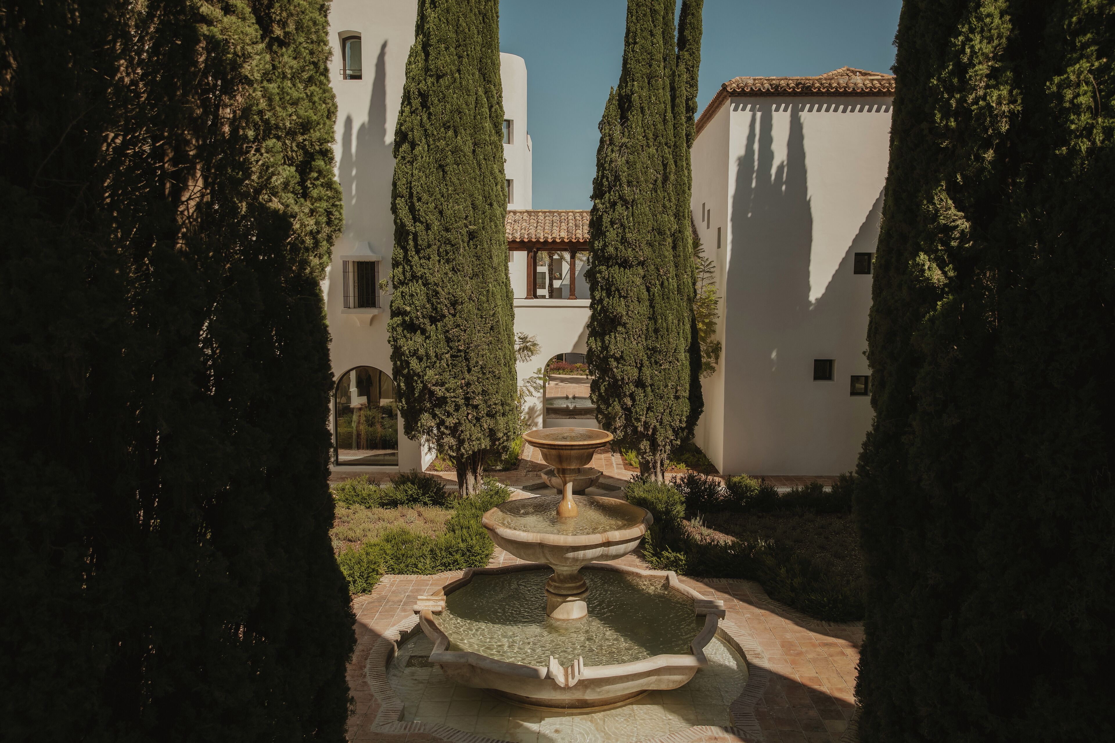 Un patio con una fuente central, rodeado de altos cipreses y un edificio con tejado de baldosas.
