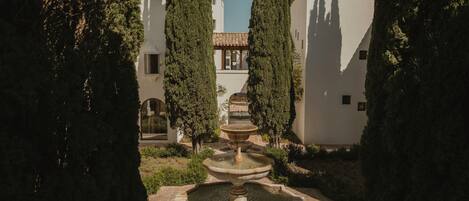 Un patio con una fuente central, rodeado de altos cipreses y un edificio con tejado de baldosas.