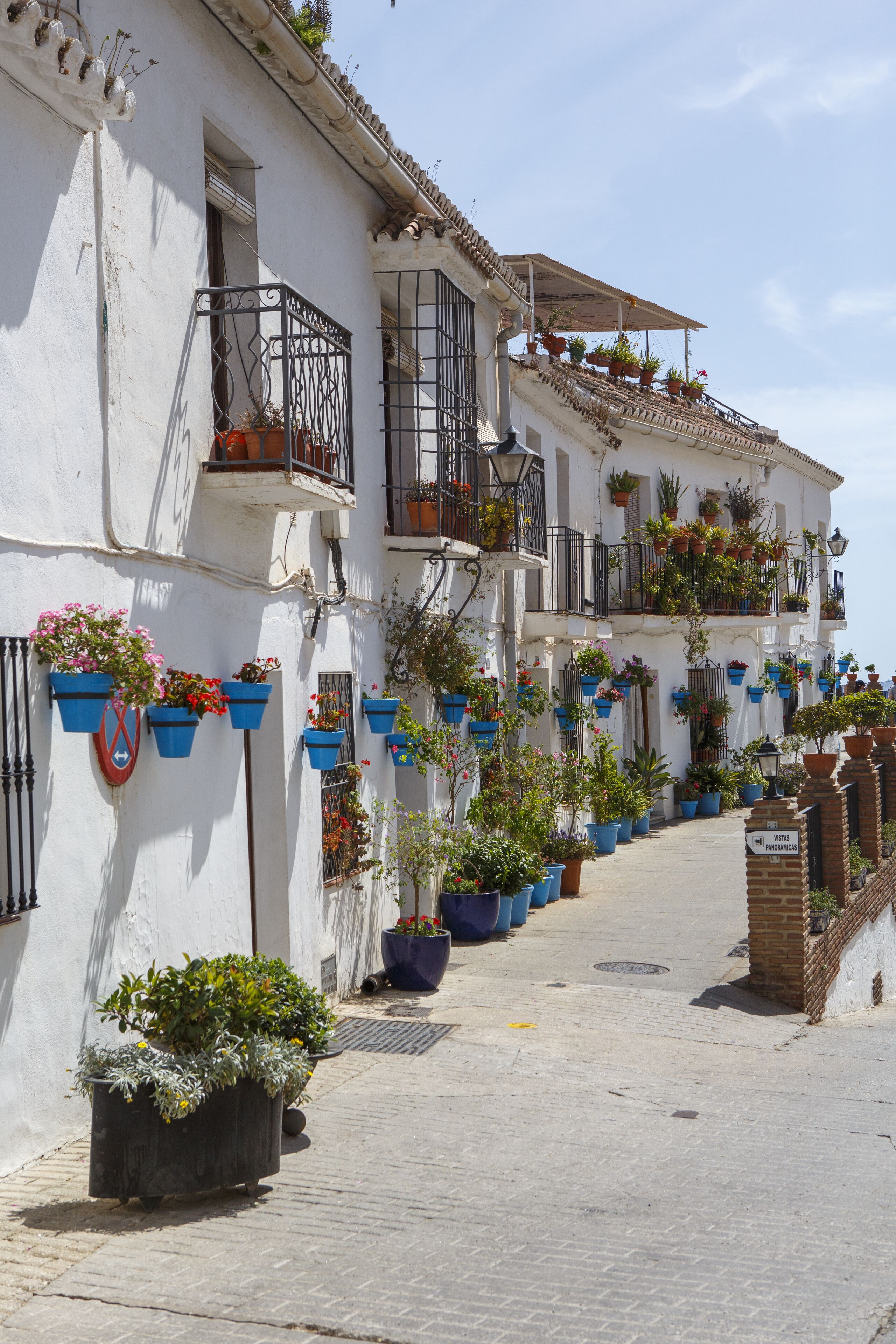 A narrow street lined with white buildings, each with balconies adorned with potted plants and flowers.