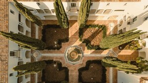 Un patio con una fuente central, rodeado de macizos de flores simétricos y altos árboles.