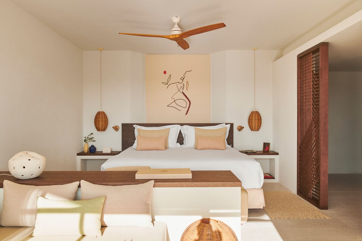 Un dormitorio con cama, mesitas de noche, ventilador de techo, una obra de arte en la pared y una cesta tejida.