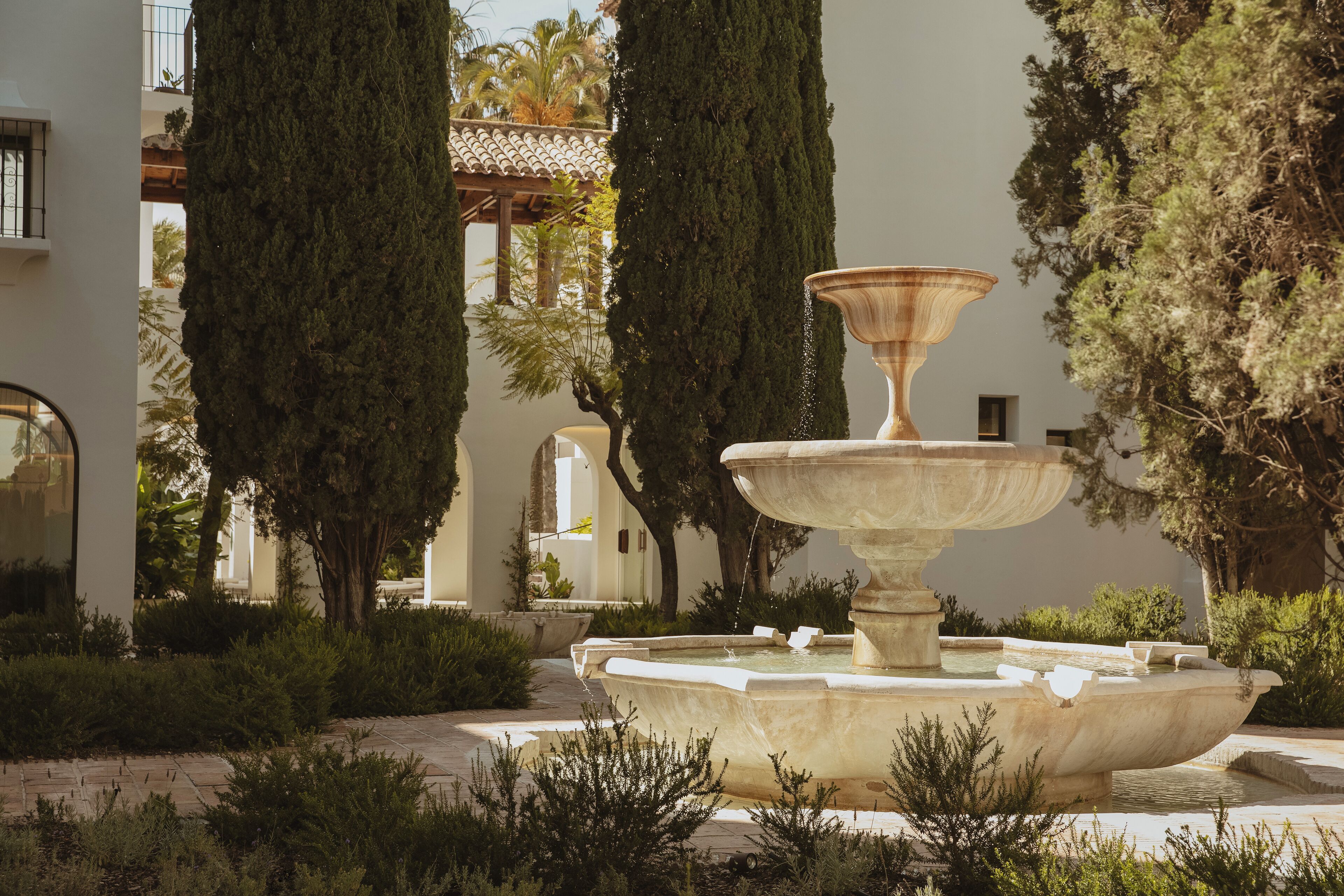 A fountain in a courtyard surrounded by tall cypress trees and a white building with arches.