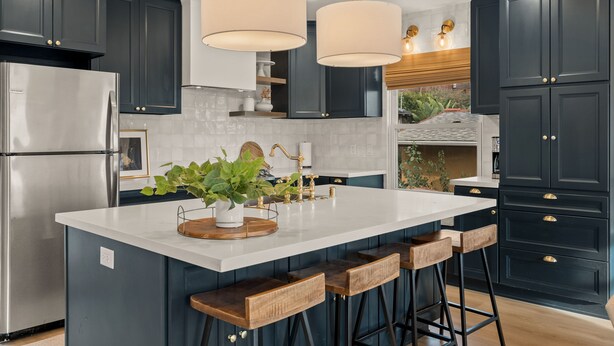 A modern kitchen with dark cabinets, a stainless steel refrigerator, a white island with bar stools, pendant lights, and a potted plant. - Modern Farmhouse in the Heart of San Diego (San Diego)