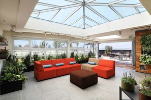 Relax in a spacious indoor area with a high ceiling and glass roof, featuring a large sofa, ottoman, and potted plants.