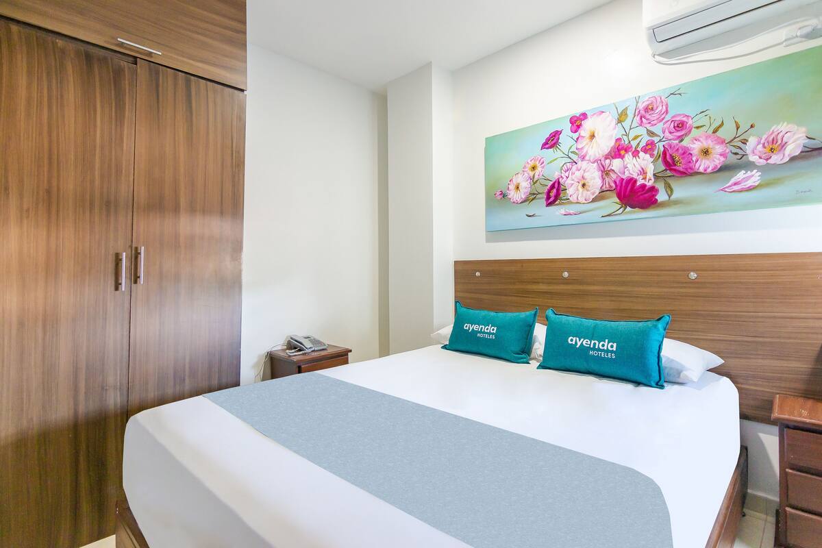 Una habitación de hotel con una cama grande, un armario de madera, una mesita de noche con un teléfono y un cuadro de flores en la pared.